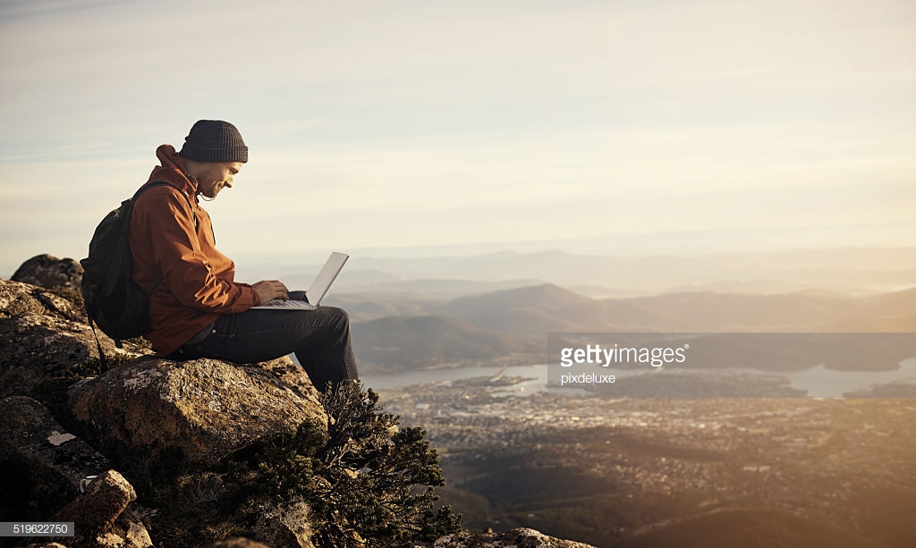 Shot of a young hiker using his laptop while sitting on top of a mountain