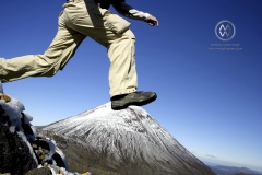 The Tongariro Crossing is considered by many to be the finest "one day walk" in New Zealand, if not the world.  The trail takes hikers through a volcanic plateau of old lava flows, volcanic craters, and gives allows the opportunity to walk between a field of three active volcanoes; Mt Tongariro, Ruapehu, and Ngauruhoe (which stars as the Lord of the Ring's Mount Doom). The ground steams with geothermal heat, and steam often rises from the active volcano cones. A hiker jumps through the air along the trail. Ngauruhoe is clearly seen in the background.